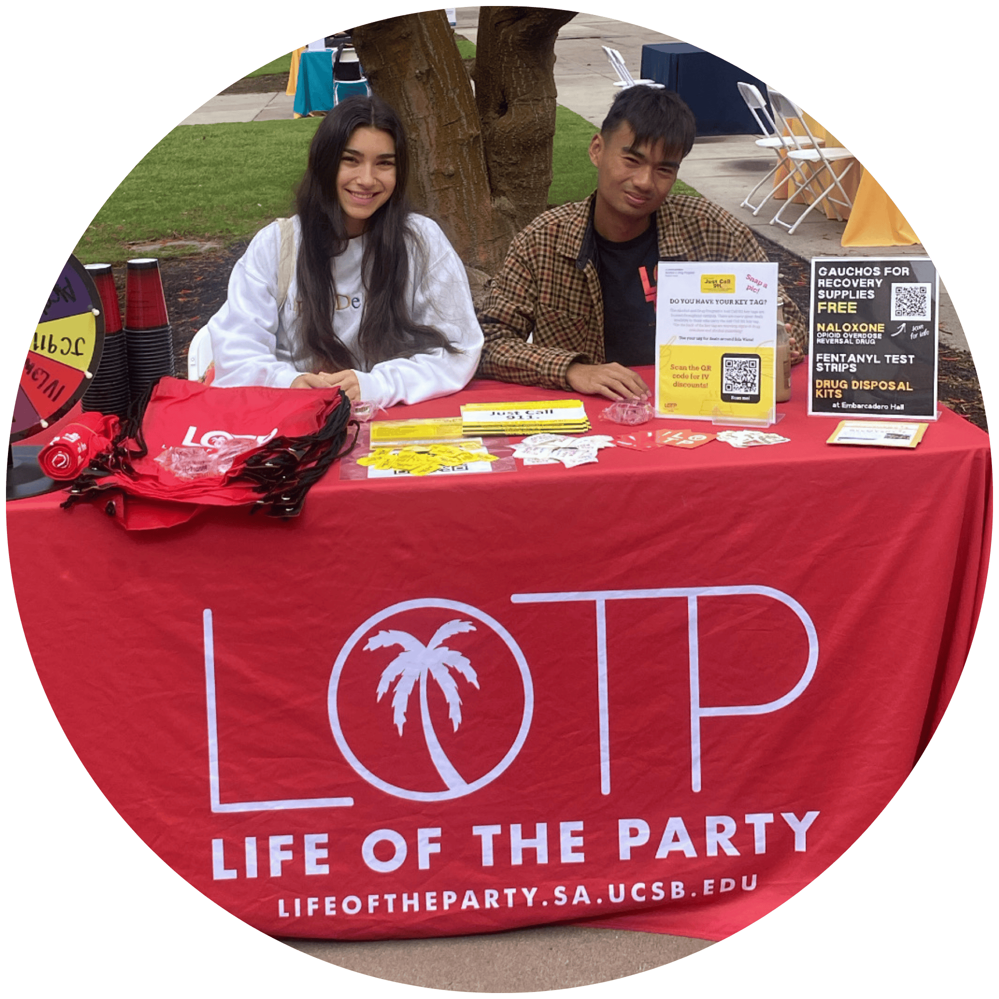Photo of 2 LOTP peers sitting at an info table with red tablecloth with the LOTP logo and literature on the table