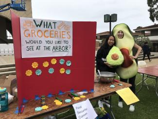 Student Holding Avocado toy next to a person dressed in an avocado costume promoting what groceries would you like to see at the arbor